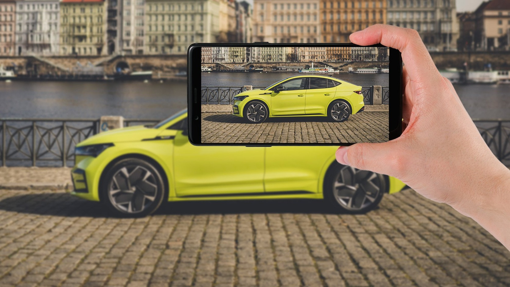 Záber na ruku s mobilom, ktorá fotí bok žltozeleného SUV. Na displeji vidno záber auta, v ktorého pozadí je mesto, budovy, rieka.